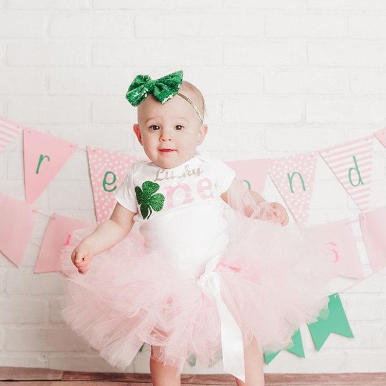 A baby girl wearing a white outfit with a green shamrock and a sparkly green bow stands in front of a white brick wall with pink and green banners.