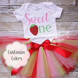 Può includere: Un body bianco a maniche corte con la scritta rosa Sweet e la scritta verde one, caratterizzato da una grafica a fragola rossa glitterata. È inclusa una gonna tutu in tulle a strati rossa, rosa e verde chiaro con un fiocco in raso rosso.