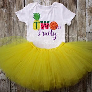 A white t-shirt with a colorful design and text, along with a yellow tulle skirt, both placed on a wooden surface.