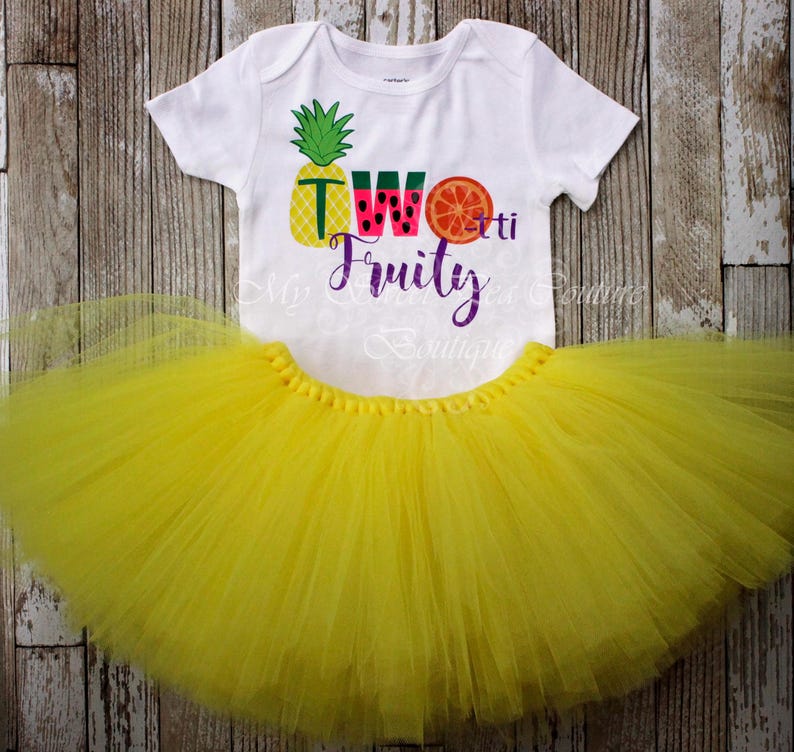 A white t-shirt with a colorful design and text, along with a yellow tulle skirt, both placed on a wooden surface.
