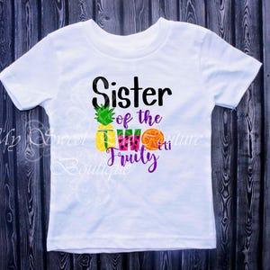 A white t-shirt with a graphic design featuring a pineapple, watermelon, and an orange, along with the text "Sister of the 2Fruit" printed on it.