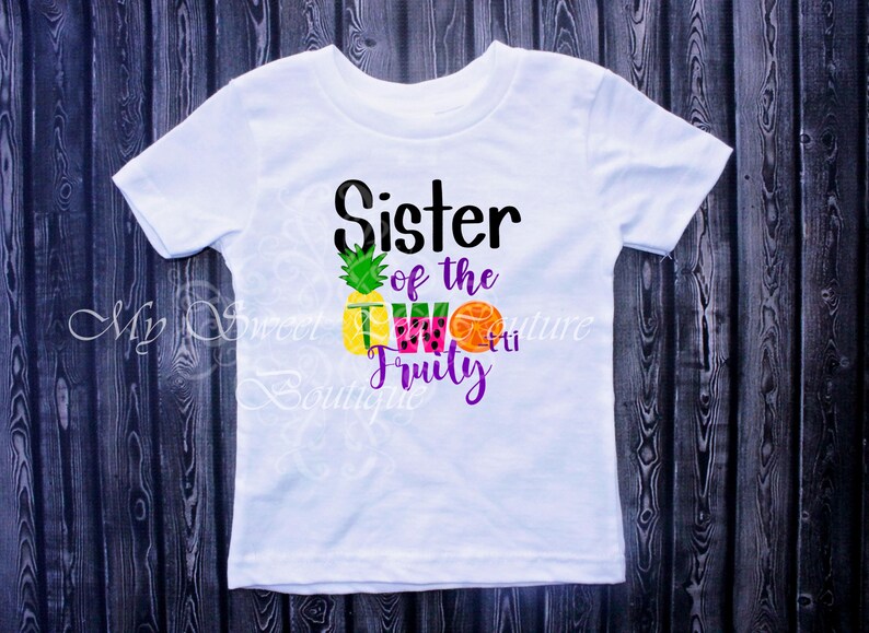 A white t-shirt with a graphic design featuring a pineapple, watermelon, and an orange, along with the text "Sister of the 2Fruit" printed on it.