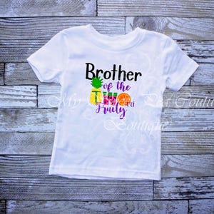 A white t-shirt with a graphic design featuring a pineapple and the text "Brother of the Two Fruits" printed on it.