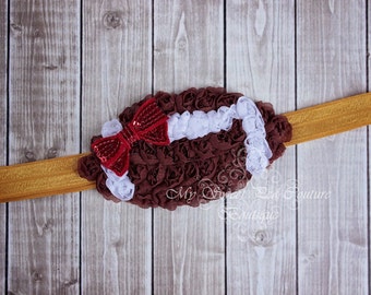 May include: A brown and white football-shaped headband with a red sequined bow. The headband is made of fabric and is decorated with ruffles. The headband is on a wooden background.