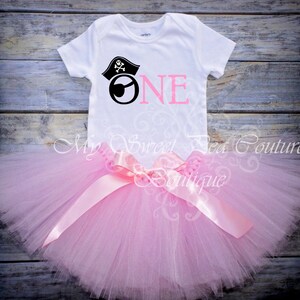 A white t-shirt with a pirate-themed design and a pink tutu skirt are displayed on a wooden surface.