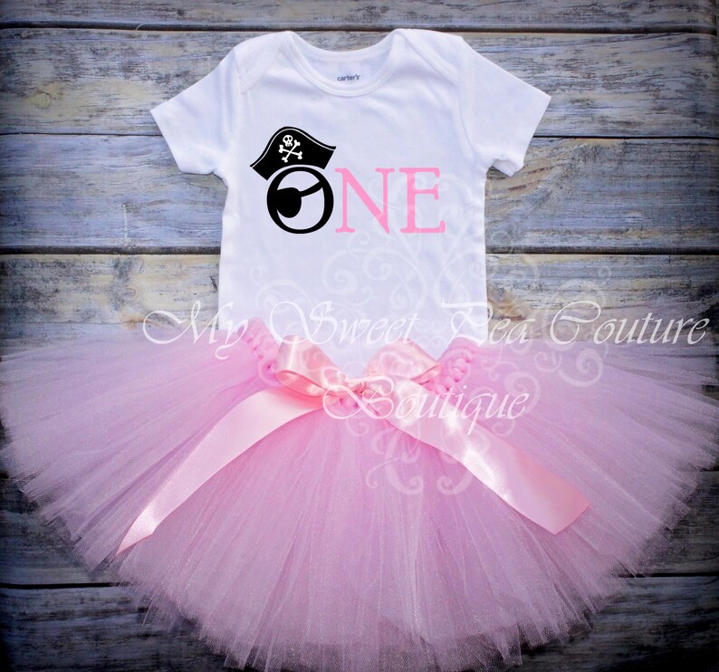 A white t-shirt with a pirate-themed design and a pink tutu skirt are displayed on a wooden surface.