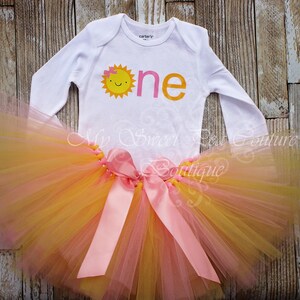 A white onesie with a sun design and a pink tutu skirt with a yellow tulle skirt are displayed on a wooden surface.