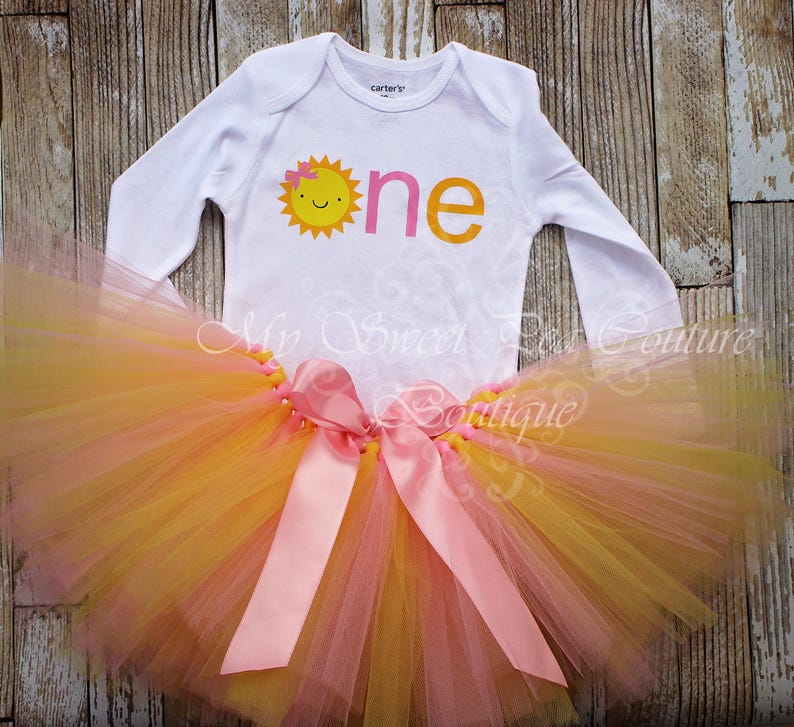 A white onesie with a sun design and a pink tutu skirt with a yellow tulle skirt are displayed on a wooden surface.