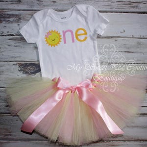 A white onesie with a sun design and a pink tutu skirt with a pink ribbon are displayed on a wooden surface.