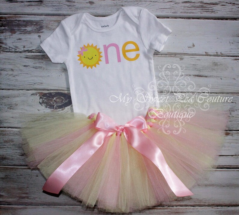 A white onesie with a sun design and a pink tutu skirt with a pink ribbon are displayed on a wooden surface.