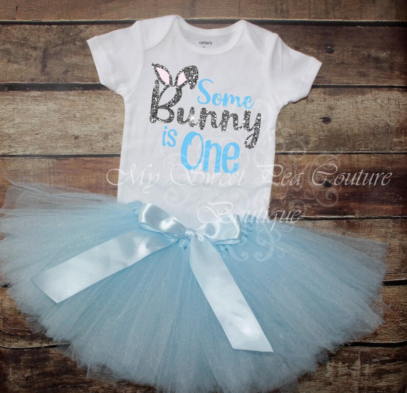 A white onesie with the text "Some Bunny is One" printed on it, along with a light blue tulle tutu skirt with a white ribbon bow.