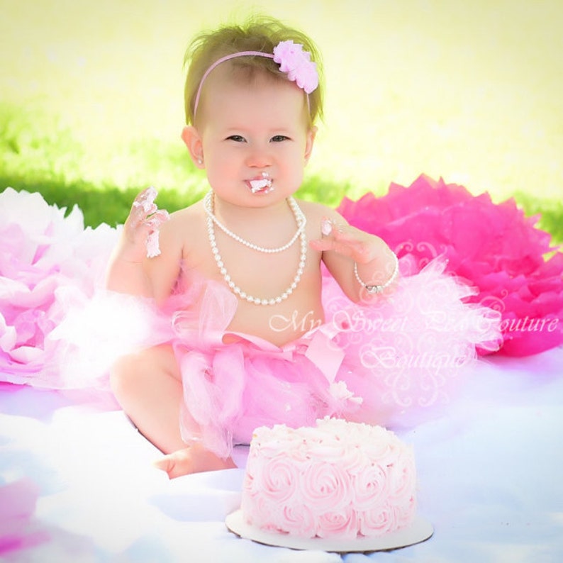 K&ouml;nnte beinhalten: Ein kleines M&auml;dchen in einem rosa Tutu und einer Perlenkette sitzt vor einem rosa und wei&szlig;en Kuchen. Sie hat Zuckerguss im Gesicht und h&auml;lt ein St&uuml;ck Kuchen in der Hand.