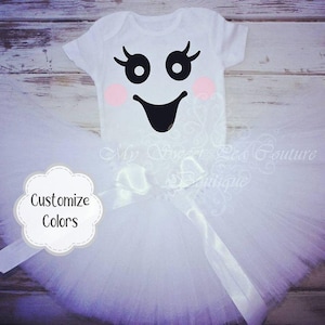 Puede incluir: Un body de bebé blanco con un diseño de cara de fantasma, combinado con un tutú de tul blanco. El body presenta ojos negros, pestañas, mejillas rosas y una sonrisa negra. Un lazo de cinta blanca adorna el tutú. El texto "Customize Colors" es visible.