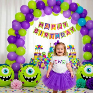 A young girl in a purple tutu and a "Happy Birthday" banner stands in front of a colorful balloon arch, with two monster-themed balloons on either side.