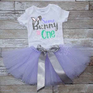 A white t-shirt with the text "Some Bunny is One" printed on it, along with a gray ribbon tied into a bow. The t-shirt is placed on a wooden surface, and a purple tulle skirt is also visible.