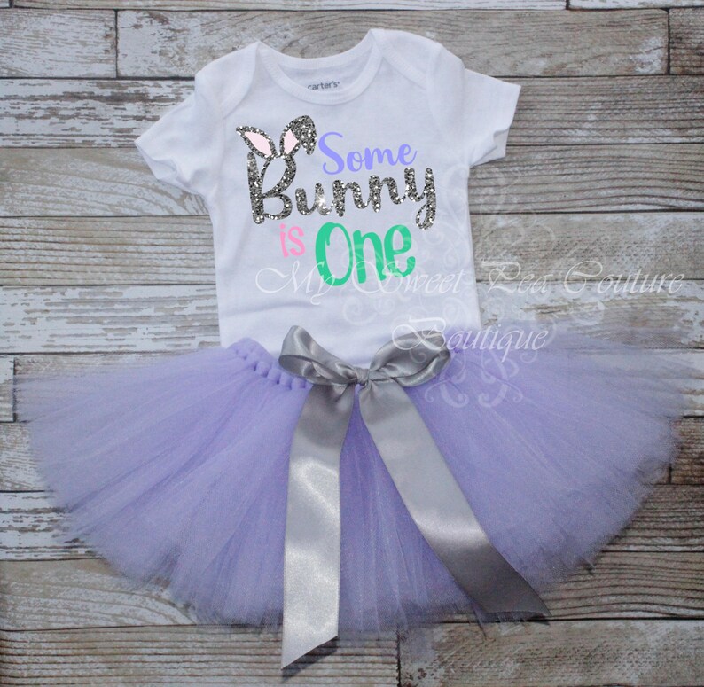 A white t-shirt with the text "Some Bunny is One" printed on it, along with a gray ribbon tied into a bow. The t-shirt is placed on a wooden surface, and a purple tulle skirt is also visible.