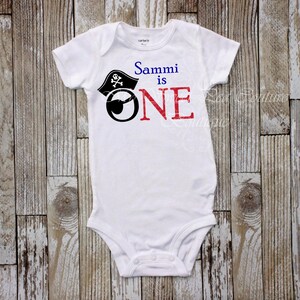 A white onesie with a pirate hat and the text "Sammi is one" printed on it, laid out on a wooden surface.