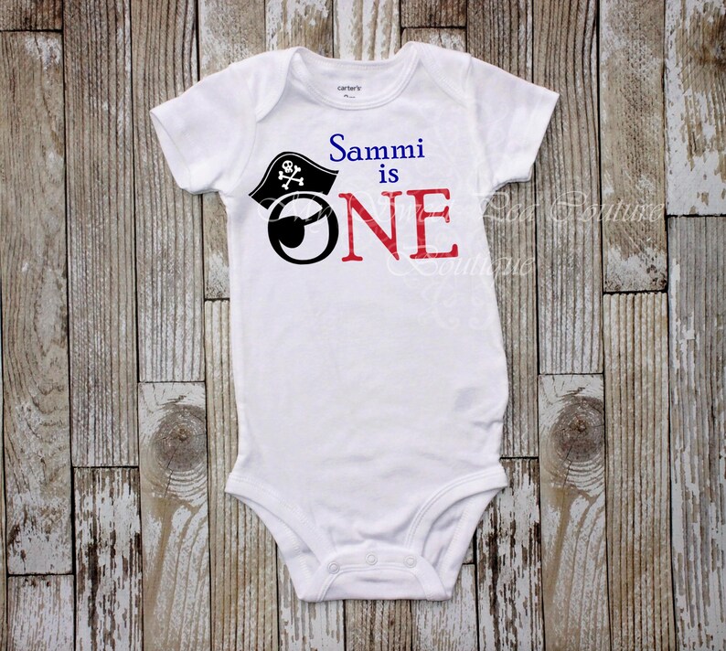 A white onesie with a pirate hat and the text "Sammi is one" printed on it, laid out on a wooden surface.