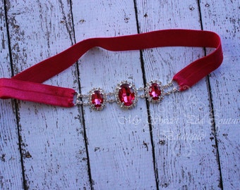 Puede incluir: Una diadema elástica rosa con tres grandes adornos de strass rojos de forma ovalada.