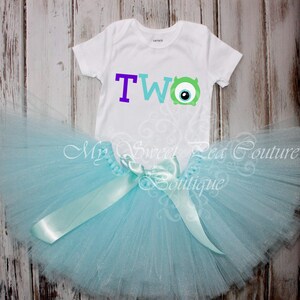 A white t-shirt with the number "TWO" and a blue monster eye design, along with a light blue tutu skirt, are displayed on a wooden surface.