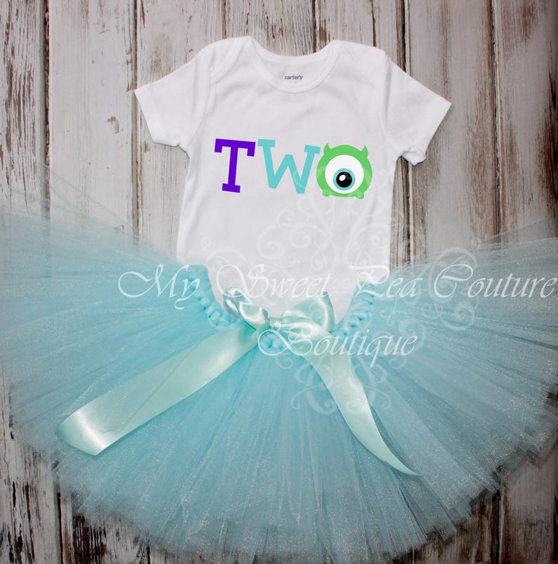 A white t-shirt with the number "TWO" and a blue monster eye design, along with a light blue tutu skirt, are displayed on a wooden surface.