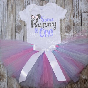 A white t-shirt with the text "Some Bunny is One" printed on it, along with a pink and purple tulle tutu skirt, laid out on a wooden surface.