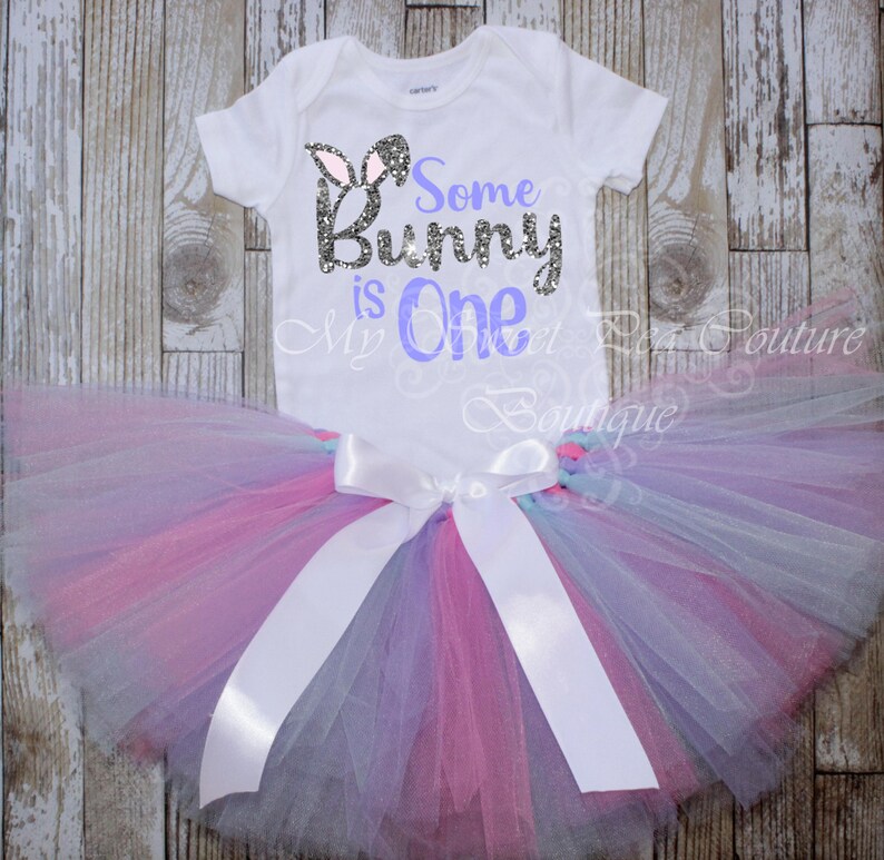 A white t-shirt with the text "Some Bunny is One" printed on it, along with a pink and purple tulle tutu skirt, laid out on a wooden surface.
