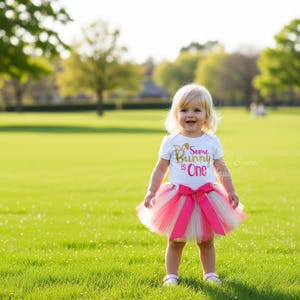 A young girl wearing a white t-shirt with the text "Some Bunny One" and a pink tutu skirt is standing in a grassy field.