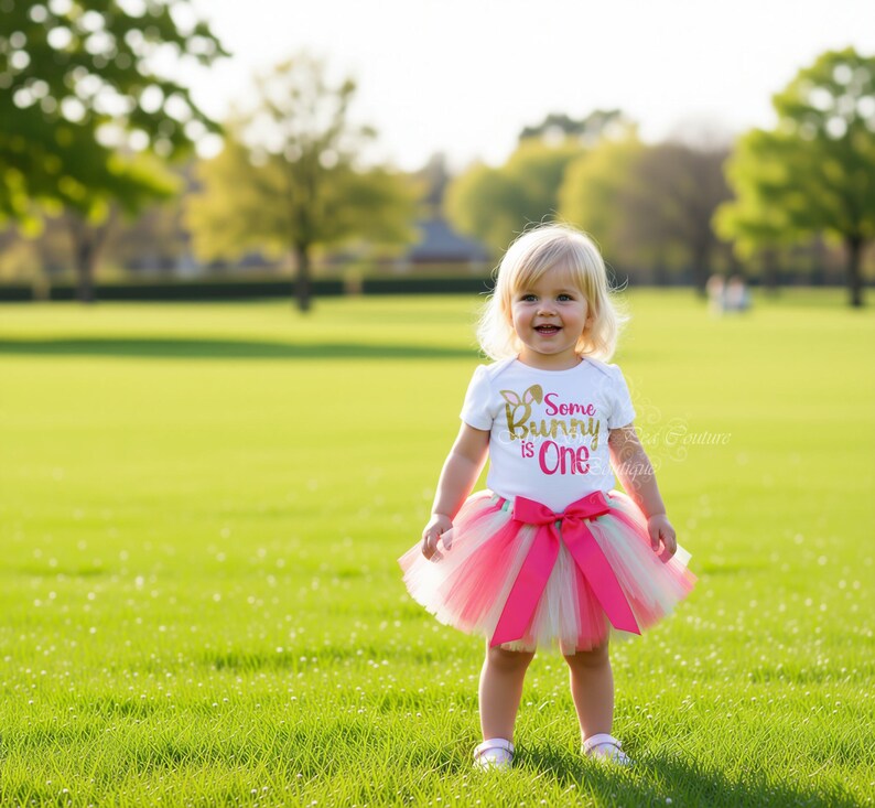 A young girl wearing a white t-shirt with the text "Some Bunny One" and a pink tutu skirt is standing in a grassy field.