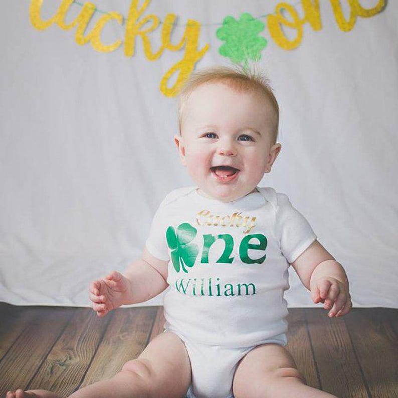 A smiling baby wearing a white onesie with a green shamrock and the name "William" printed on it, sitting on a wooden floor in front of a banner with the word "lucky" on it.