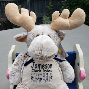 May include: A soft toy moose with large antlers, embroidered with the name "Jameson" and birth details. The moose is light brown with a cream-colored belly and is sitting on a blue and white rocking chair. The text includes the baby's birth time, weight, and length (48 cm).