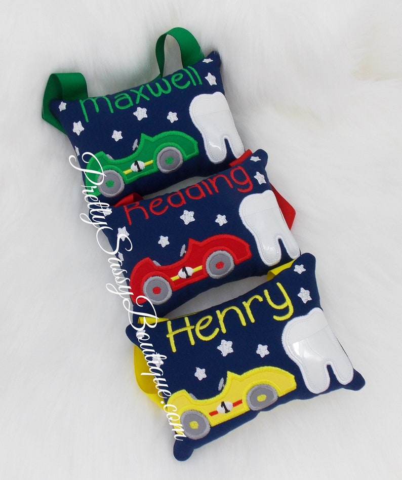May include: Three blue tooth fairy pillows with white teeth, each with a different colored race car and a name embroidered on the pillow: Maxwell, Reading, and Henry.