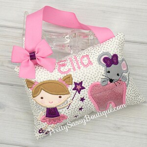 May include: A white pillow with a silver glitter polka dot pattern. The pillow features a pink tooth, a gray mouse with a purple bow, a girl with brown hair and a purple dress, and a purple star. The pillow is personalized with the name "Ella" and the text "Pretty Sassy Boutique.com".