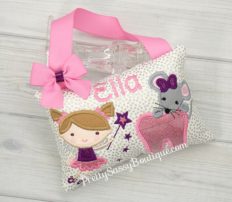 May include: A white pillow with a silver glitter polka dot pattern. The pillow features a pink tooth, a gray mouse with a purple bow, a girl with brown hair and a purple dress, and a purple star. The pillow is personalized with the name "Ella" and the text "Pretty Sassy Boutique.com".