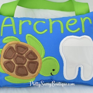 May include: A blue pillow with a green embroidered turtle and a white tooth pocket. The pillow has the name "Archer" embroidered on it in green.