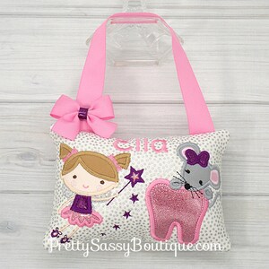 May include: A white pillow with a silver glitter pattern, featuring a tooth fairy with a wand, a mouse holding a tooth, and the name "Ella". The pillow has pink ribbon straps and a pink bow.