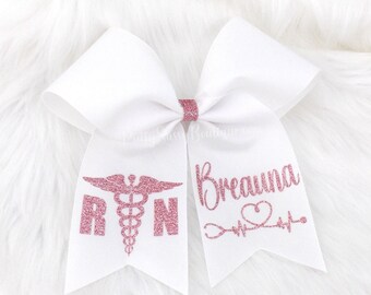 May include: White cheer bow with pink glitter lettering. The left side of the bow says "RN" with a pink glitter caduceus symbol. The right side of the bow says "Breanna" with a pink glitter stethoscope, heart, and EKG line.