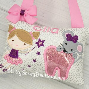 May include: A white pillow with a gray mouse holding a pink tooth, a girl fairy with a wand, and the name "Ella" embroidered on it. The pillow has a pink ribbon loop for hanging.