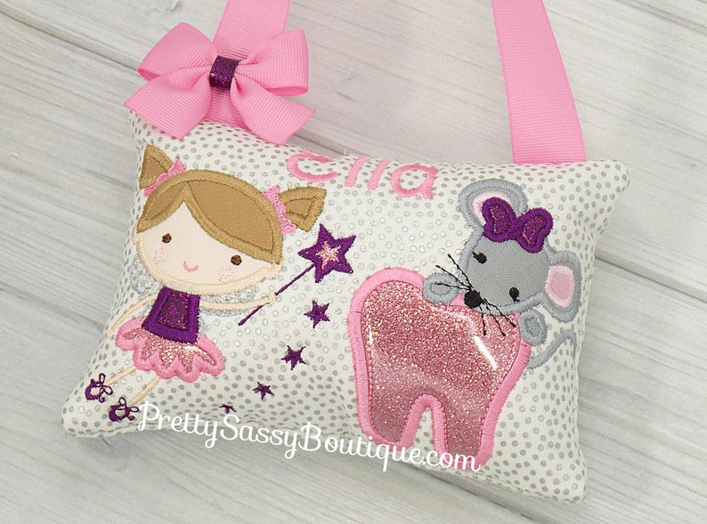 May include: A white pillow with a gray mouse holding a pink tooth, a girl fairy with a wand, and the name "Ella" embroidered on it. The pillow has a pink ribbon loop for hanging.