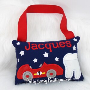 May include: A blue tooth fairy pillow with a red race car and white stars. The pillow has a red ribbon loop and the name "Jacques" embroidered on it.