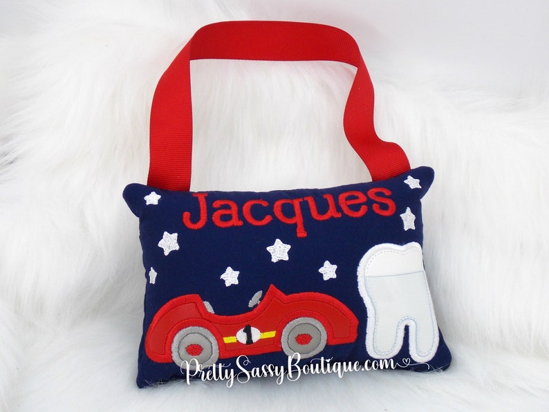 May include: A blue tooth fairy pillow with a red race car and white stars. The pillow has a red ribbon loop and the name "Jacques" embroidered on it.
