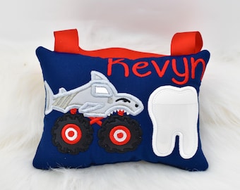 Könnte beinhalten: Ein blaues Zahnkissen mit einem Hai-Monstertruck-Design und einer weißen Zahntasche. Das Kissen hat rote Bänder und der Name "Kevyn" ist darauf gestickt.