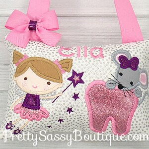 May include: A white pillow with a silver glitter background and a pink tooth, a purple fairy with a wand, and a gray mouse with a purple bow. The pillow has a pink ribbon loop and the name "Ella" is embroidered on the pillow.