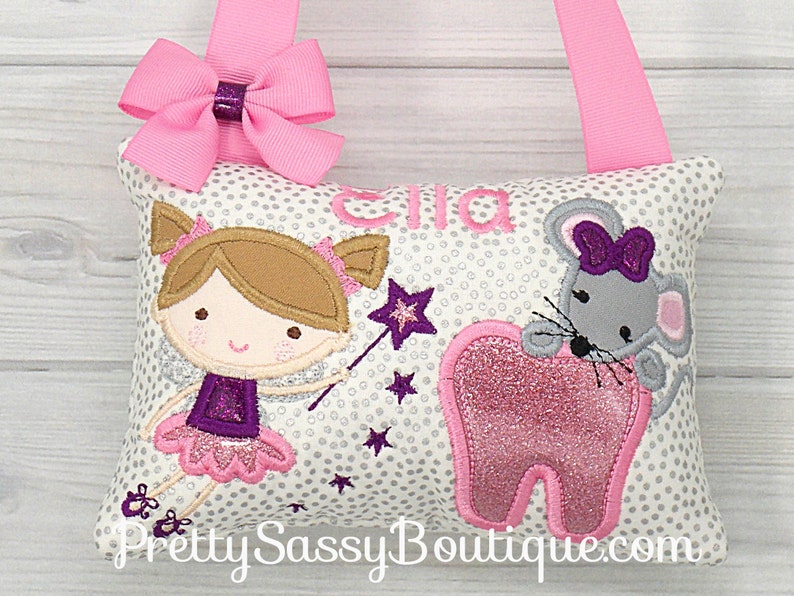 May include: A white pillow with a silver glitter background and a pink tooth, a purple fairy with a wand, and a gray mouse with a purple bow. The pillow has a pink ribbon loop and the name "Ella" is embroidered on the pillow.
