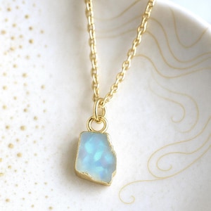May include: A gold-colored chain necklace with a pendant. The pendant is a light blue, irregular-shaped stone with a gold-colored border. The necklace is displayed on a white surface with gold-colored decorative lines.