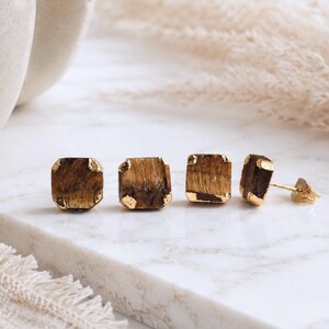 Pode incluir: Quatro brincos de tarracha quadrados com pedras preciosas olho-de-tigre polidas em armações de metal dourado. Cada brinco tem uma superfície castanha e dourada chatoyante com grão visível e um pino dourado para orelhas furadas.