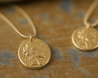 May include: Two gold-colored coin necklaces. Each pendant features a profile of a person with detailed hair and a chain. The necklaces are displayed on a textured, brown and blue surface. The pendants are round and have a shiny finish.