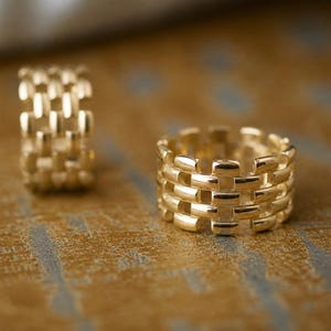 May include: Two gold-colored rings with a woven design. The rings are made of a shiny metal and are displayed on a textured, golden-brown surface. The rings are similar in style, with one ring in focus.