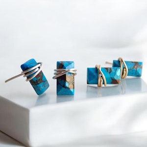 May include: Four pairs of turquoise and brown stone earrings. The rectangular stones are wrapped with silver or gold wire. The earrings are displayed on a white surface.