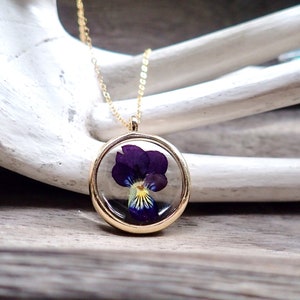 May include: A gold chain necklace with a round pendant featuring a preserved purple pansy flower.
